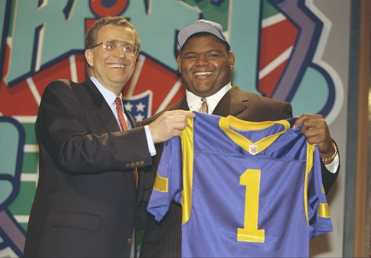 1997 — ORLANDO PACE
<b>St. Louis Rams</b> | T Ohio State
<br>El liniero ofensivo Orlando Pace sostiene su nueva camiseta de los St. Louis Rams frente a su nuevo armario en las instalaciones de prácticas de los Rams el sábado 19 de abril de 1997 en St. Louis. Pace de Ohio State, fue la primera selección general en el draft de la NFL. (Foto AP / Tom Gannam)